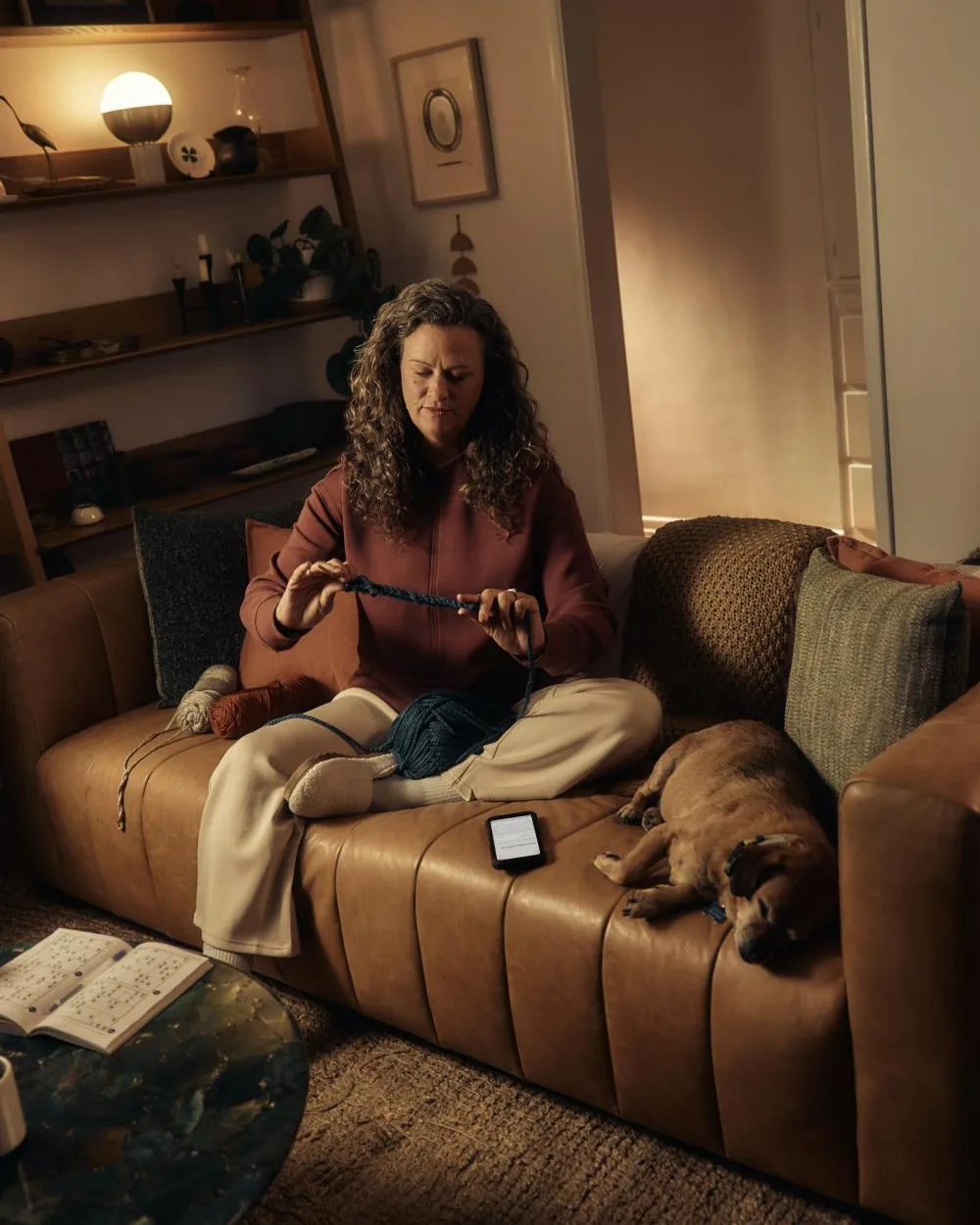 A woman knitting on a leather sofa in a warmly lit living room, her dog asleep beside her