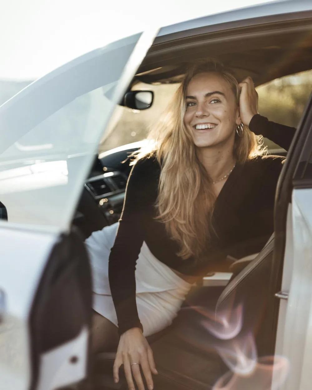 A smiling young woman sitting in the driver’s seat of a car with the door open