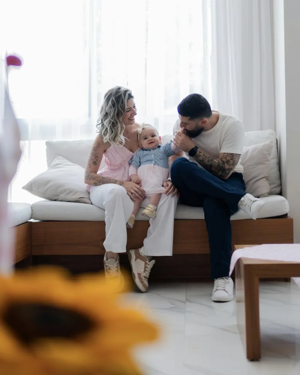 A mother and father on a sofa in a bright living room, laughing with their baby daughter