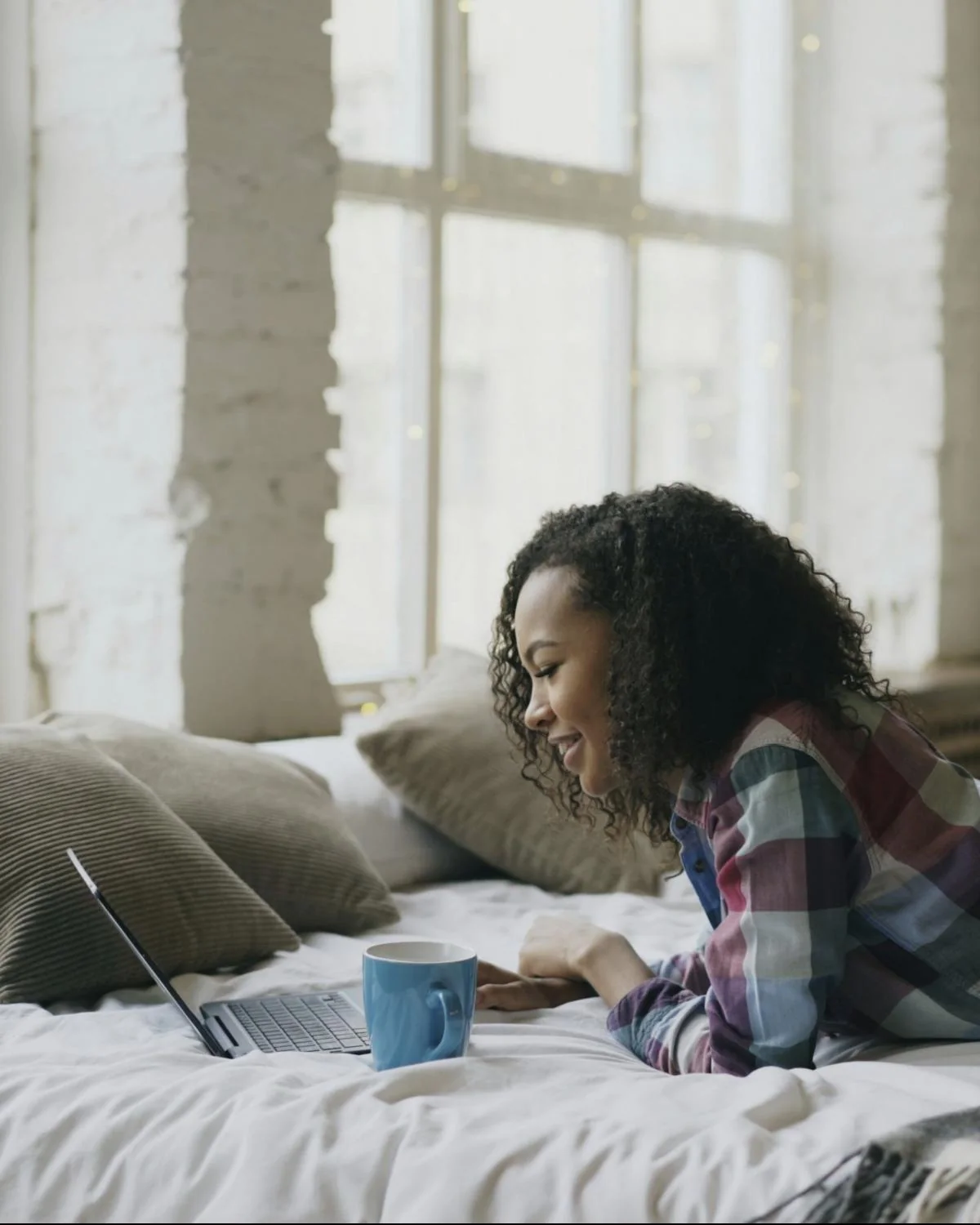 A young woman lying on her bed, smiling at her laptop with a cup of coffee